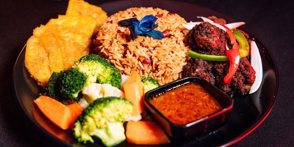 A vibrant plate of rice, fried plantains, steamed vegetables, meatballs, and sauce.