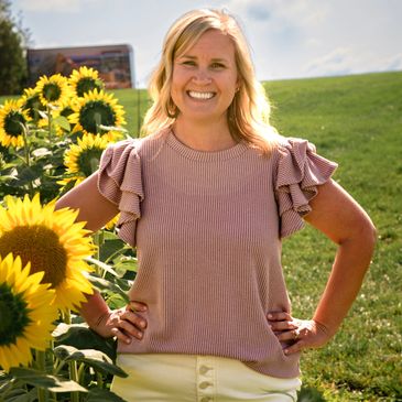 Smiling woman standing confidently among sunflowers on a sunny day.