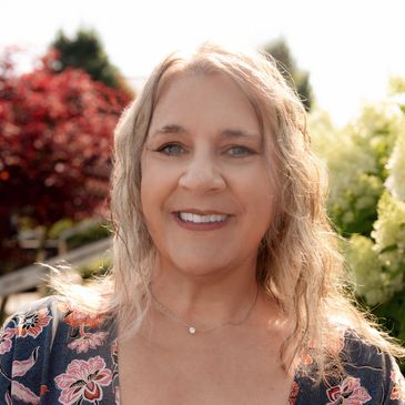 Smiling woman in floral dress stands outdoors with greenery and sunlight.