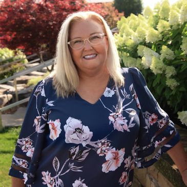 Smiling woman in floral blouse stands outdoors by flowers and stone wall.