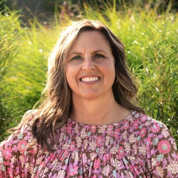 Smiling woman in a floral dress stands outdoors with greenery behind her.