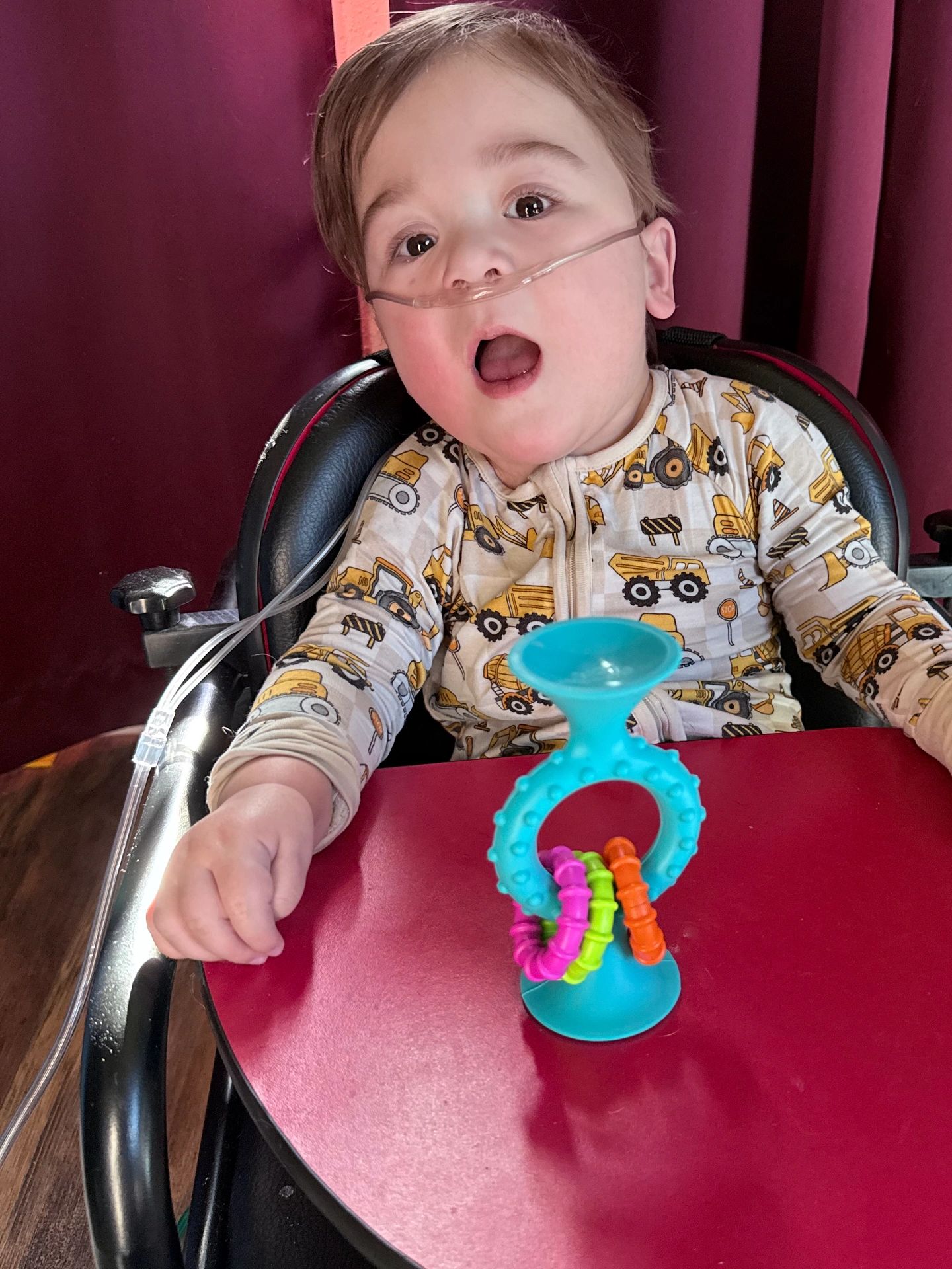 A toddler with medical oxygen tubes sits in a high chair playing with a colorful toy.