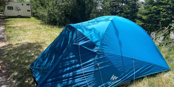 A blue Quechua tent pitched on grass near trees and a caravan.