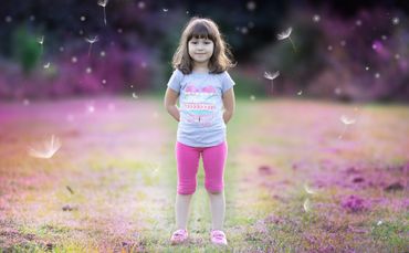 A young girl stands in a field with dandelion seeds floating around her.