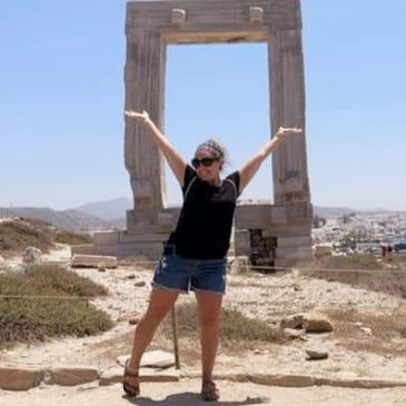Woman posing joyfully in front of an ancient stone gateway under a clear blue sky.