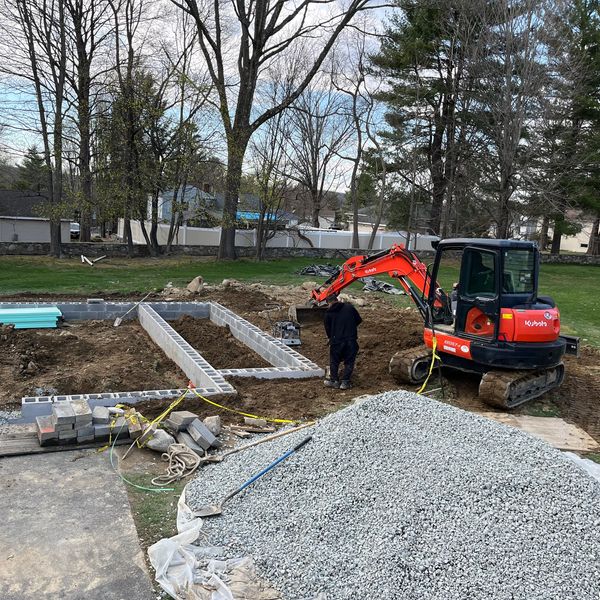 Construction site with a worker operating a small excavator near foundation blocks.