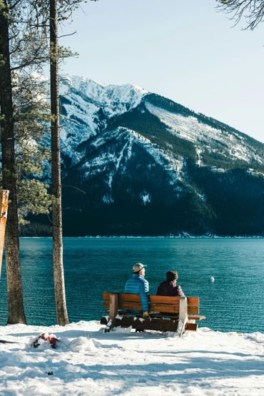Two people sitting on a bench by a snowy lake with mountain views.