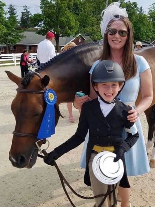 Young equestrian with a horse wearing a blue ribbon, posing with a woman outdoors.