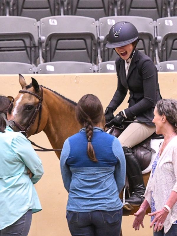 A joyful equestrian on horseback shares a laugh with three onlookers indoors.