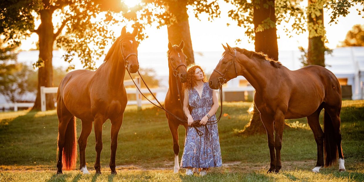 A woman stands with three horses in a sunlit park during golden hour.
