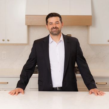 Man in black suit standing confidently in a modern white kitchen.
