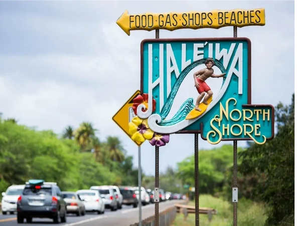 Colorful Haleiwa North Shore sign with surfing theme and traffic in background.