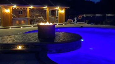 Cozy nighttime pool area with fire pit and illuminated blue water.