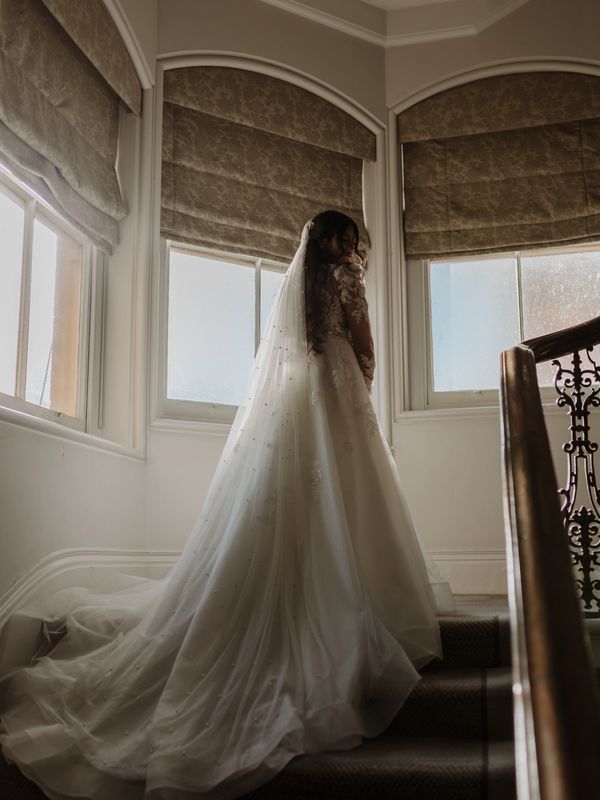 A bride wearing a white wedding dress and veil stands on a staircase.