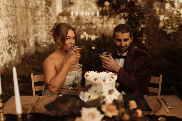 A bride and a groom sit at a table and sip from cocktail glasses. On the table is a white cake.