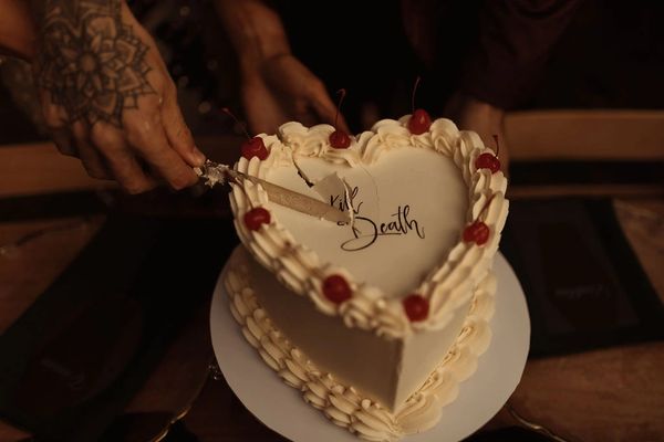 A white heart shaped wedding cake, decorated with red cherries and 'Til Death' in black script.