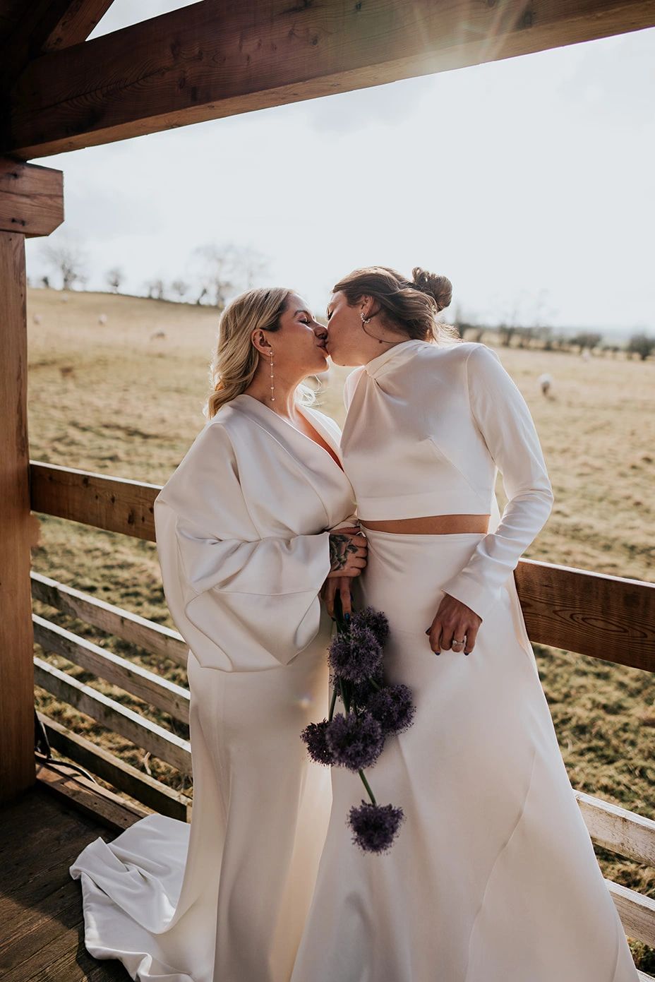 Two brides wearing white gowns kiss. One holds a bouquet of purple alliums in her hand.