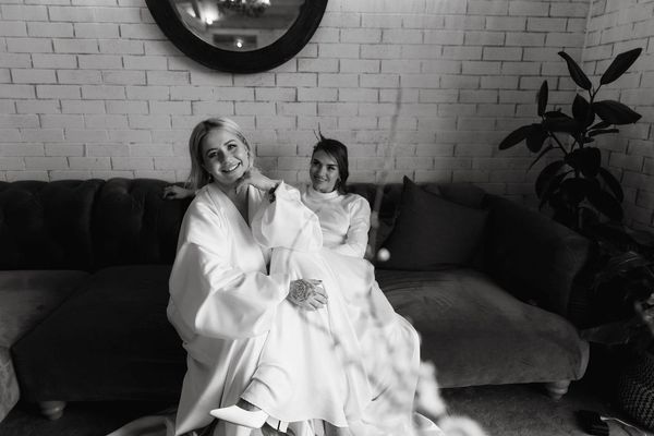 Two brides wearing white gowns sit on a sofa. One leans forward with her hand under her chin.