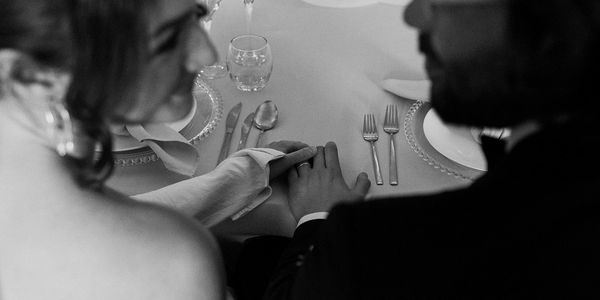 A bride and groom are photographed seated at a table, holding hands.