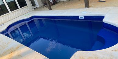 A clean blue swimming pool next to a patio with chairs and a broom.