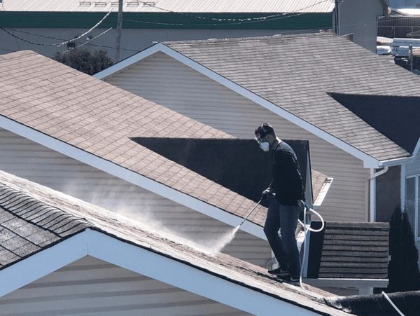A person power washing a house roof while wearing a protective mask.