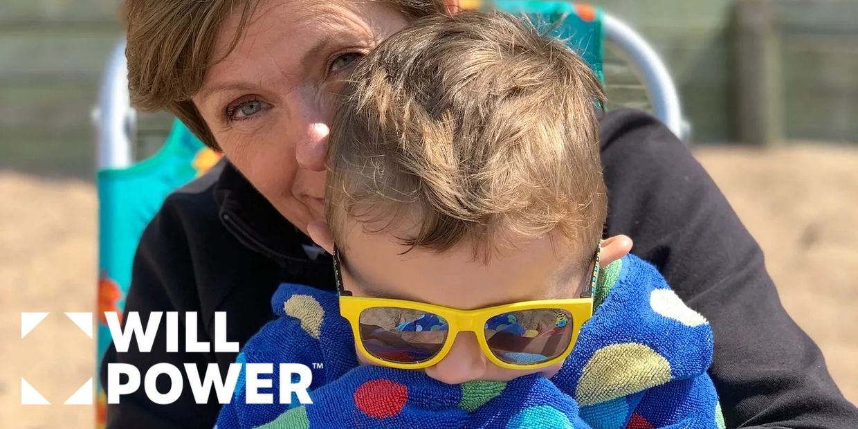 A woman hugs a child wrapped in a colorful towel with yellow sunglasses at the beach.