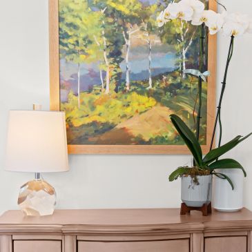 Lamp, orchid, and landscape painting on wooden dresser.