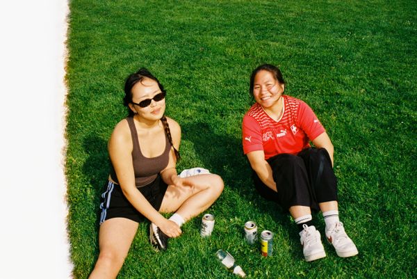 Two women sitting on grass, enjoying drinks on a sunny day.