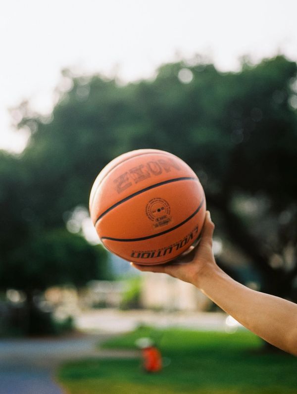 A hand holding a basketball outdoors with blurred trees in the background.