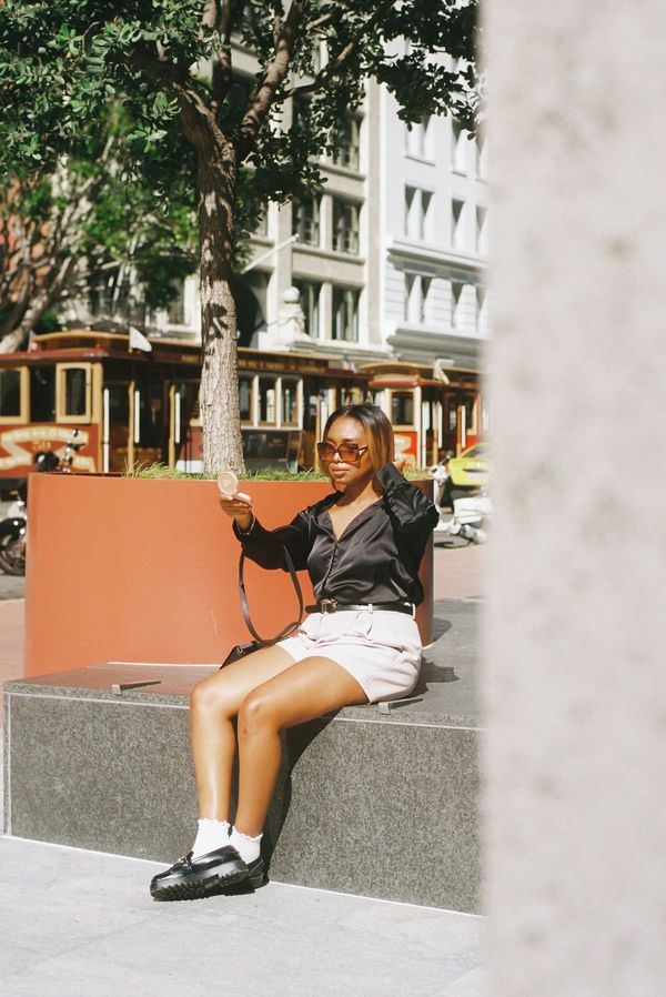 Woman in sunglasses sitting outdoors, holding a compact mirror.