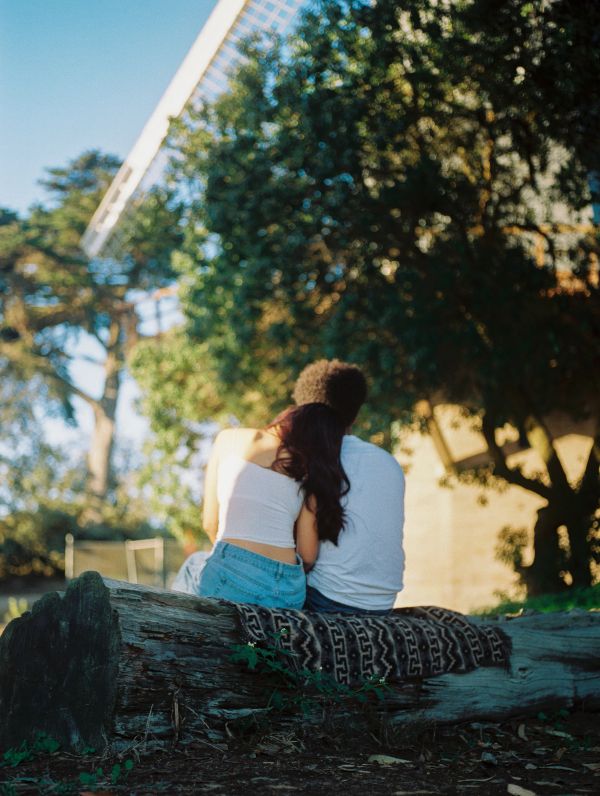 A couple sitting closely on a blanket-covered log in a sunny park.