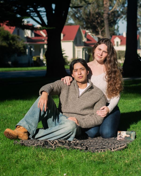 A couple sitting on a blanket in the sun on green grass.