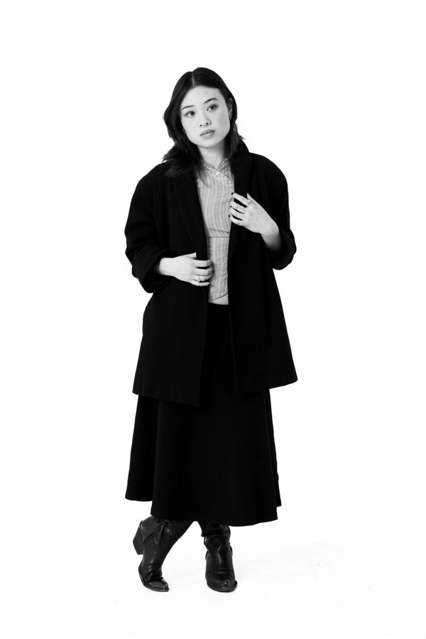 Woman in black coat and boots posing thoughtfully against white background.