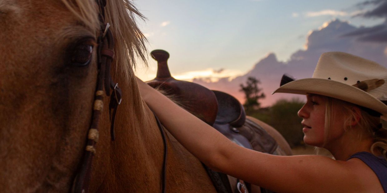 A girl in a cowboy hat pets a horse at sunset.