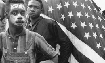 Two young men stand in front of an American flag, one with 'VOTE' painted on his forehead.