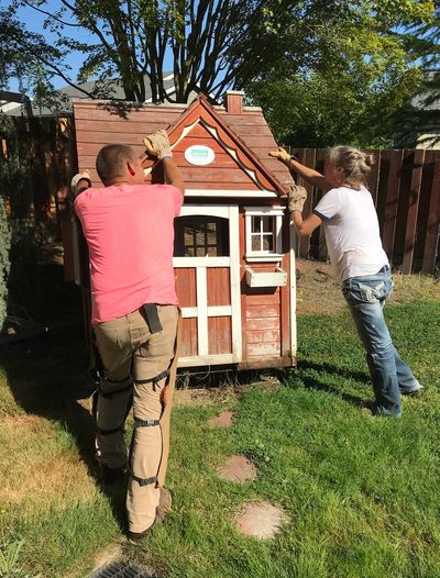 Two people repairing a small wooden playhouse outdoors.