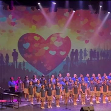 Children's choir singing on stage with a heart backdrop.