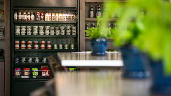 Shelves stocked with various packaged food items behind a blurred dining area with green potted plants.