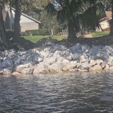 Rip rap shoreline with trees and houses in the background by a calm water body.