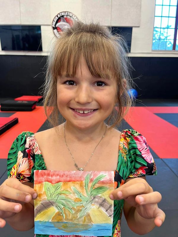 Smiling girl proudly shows her colorful palm tree painting indoors.