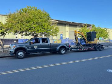 Pickup truck towing a trailer with a compact excavator on an urban street.