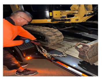 Man securing a construction vehicle with chains on a trailer at night.
