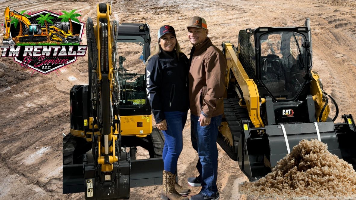 A smiling couple stands between heavy construction equipment on a dirt site.