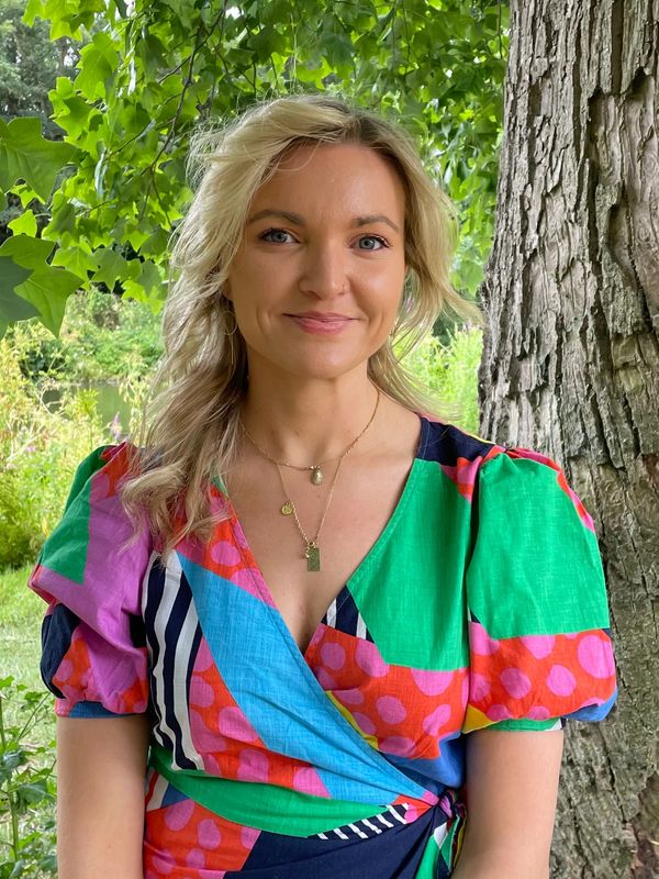 Smiling woman in colorful dress standing by a tree outdoors.