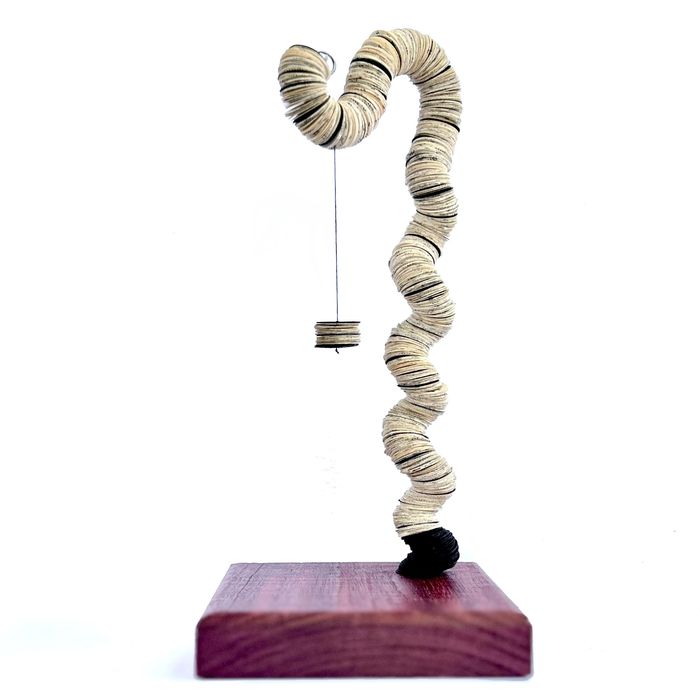 A winding paper form suspended above a wood base with a small book page element hanging by a thread.