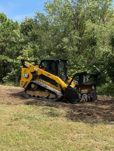 Yellow CAT compact track loader parked on cleared ground near green trees.