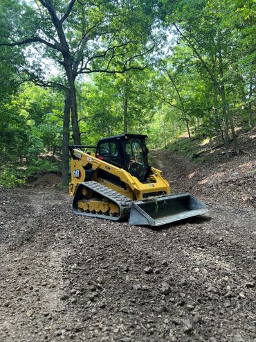 A yellow CAT skid steer loader on a dirt path in a forest.