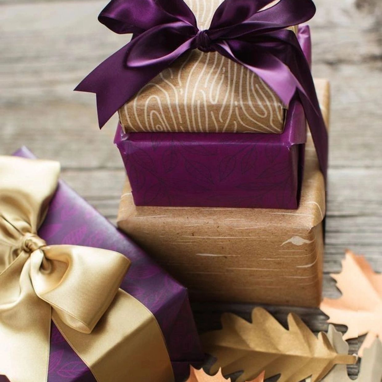 Stacked gift boxes with purple and gold ribbons surrounded by autumn leaves.
