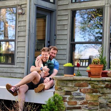 Husbands Todd behind Rob hugging and smiling on mountain house front porch in spring