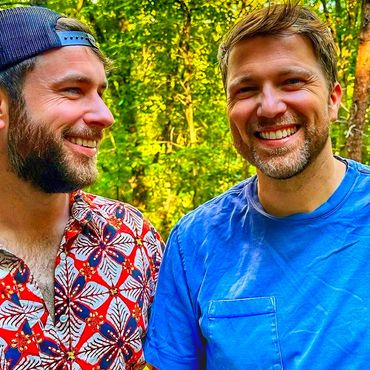 Husbands Todd with hat backward, smiling at Rob with the summer forest and leaves in the background